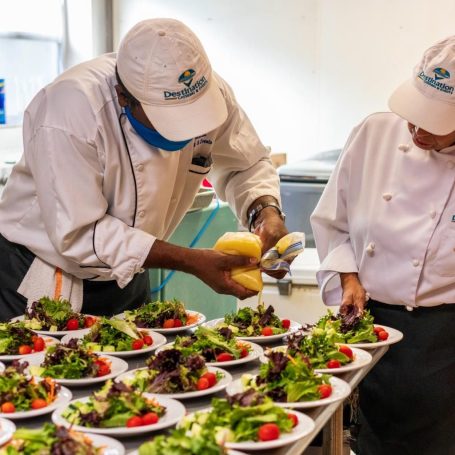 Destination Catering Private Chefs at sea. Two chefs plating salads with fresh greens and cherry tomatoes in a kitchen setting.