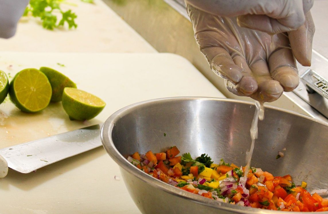 Mango Salsa Preparation. A chef squeezes lime juice over a colorful mixed salad in a stainless steel bowl.
