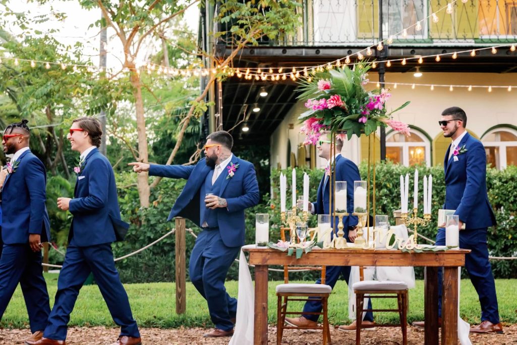 Party Time Groomsmen in navy suits walking past a decorated table at an outdoor wedding.