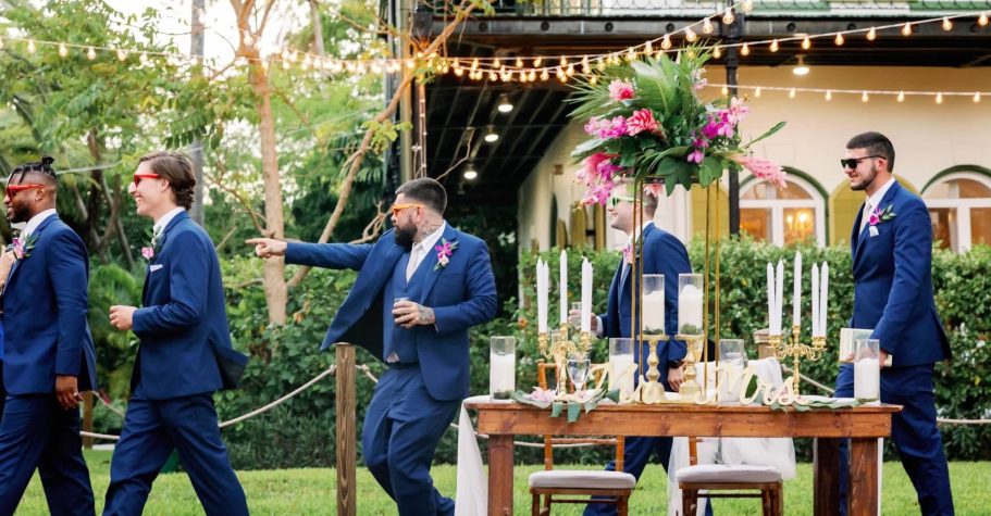 Groomsmen in blue suits walking past a floral decorated table at an outdoor wedding.