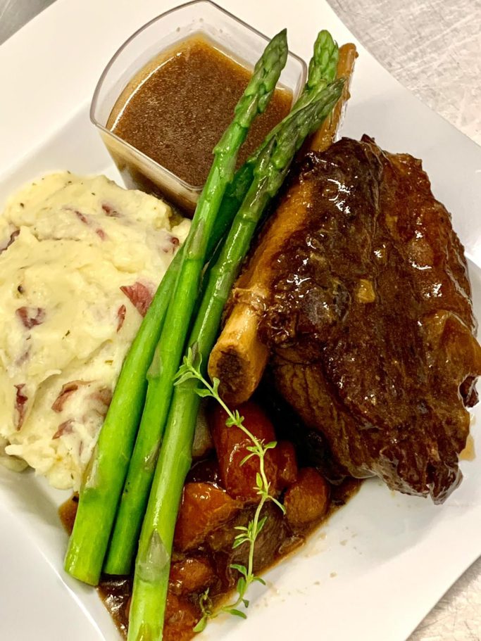 Braised Short Rib, herbed-creamed potato A plate featuring braised meat, mashed potatoes, asparagus, and a sauce.
