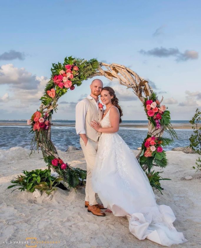 Beach Wedding A couple in wedding attire stands together by a floral arch on a beach.