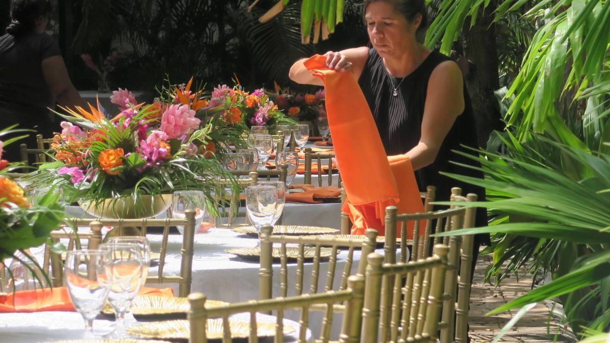 Key West Garden Reception A woman arranging orange napkins on a beautifully set outdoor dining table with flowers.