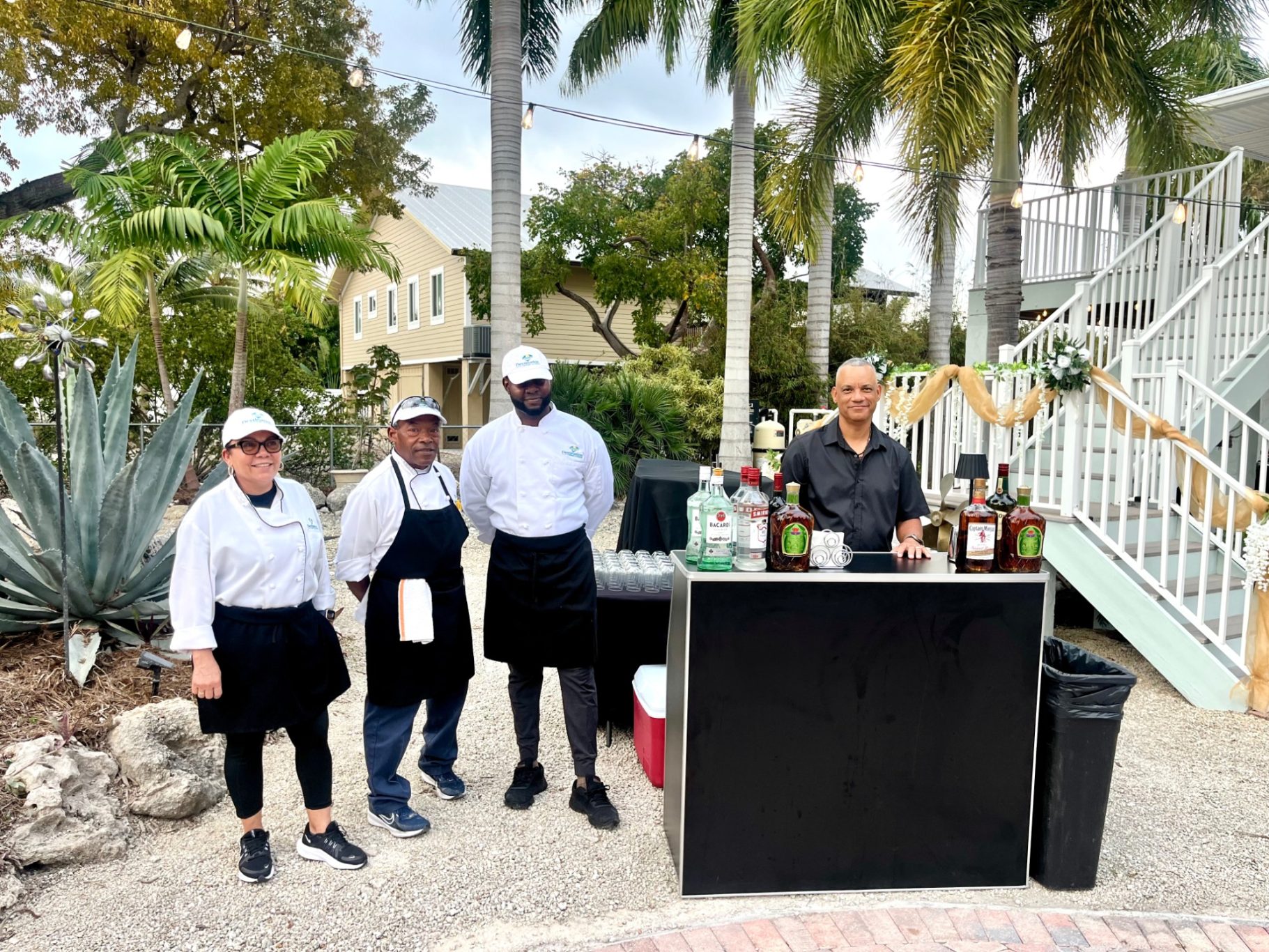 Event Crew Four chefs stand outside near a bar with various bottles, surrounded by greenery.