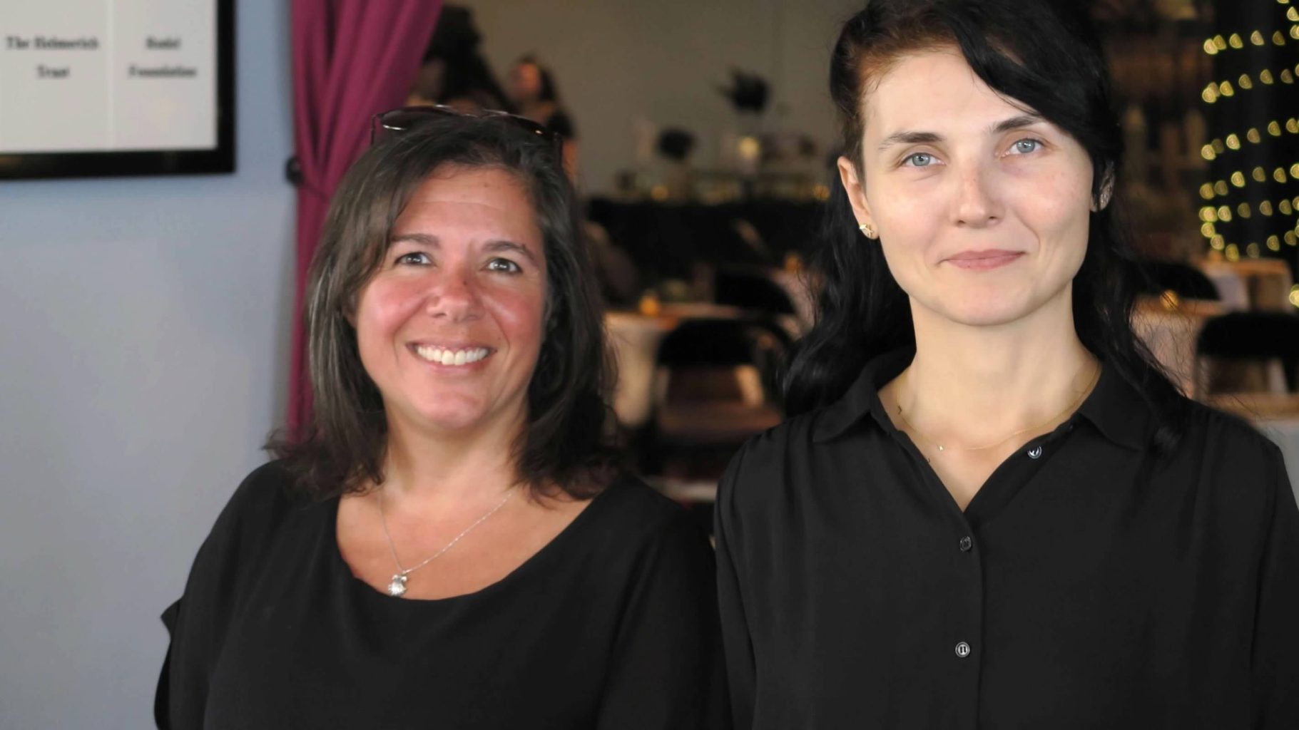 Catering Crew Team Two women smiling, wearing black tops, standing together in a dining setting.