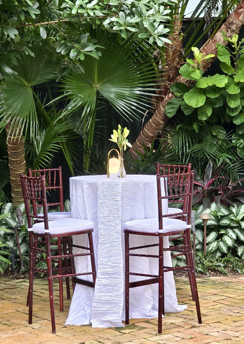 Pool Side-Garden Cocktail Reception A round table with a white tablecloth and four high chairs surrounded by lush greenery.