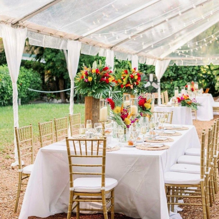 Elegant outdoor dining setup under a clear tent with floral centerpieces and white tablecloths.