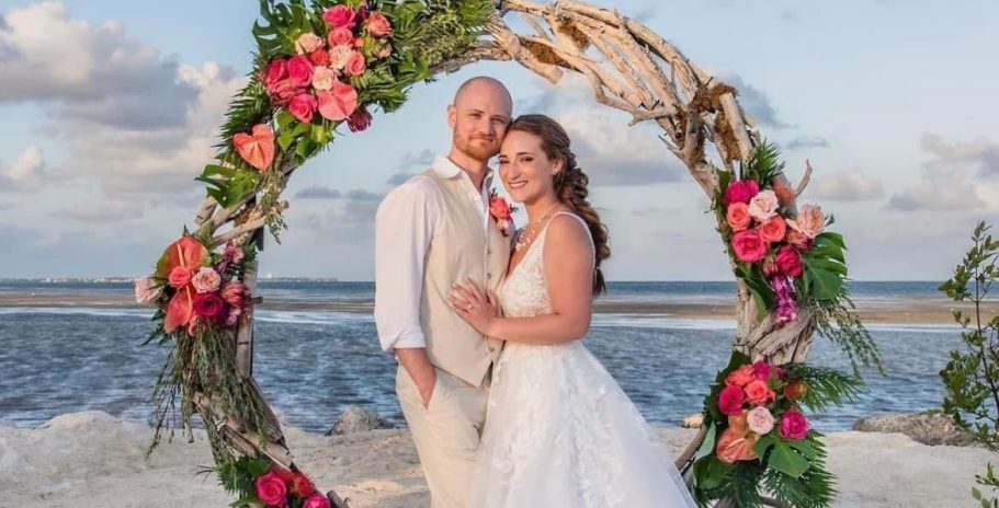 Bride and groom pose together in a floral arch on a beach at sunset.